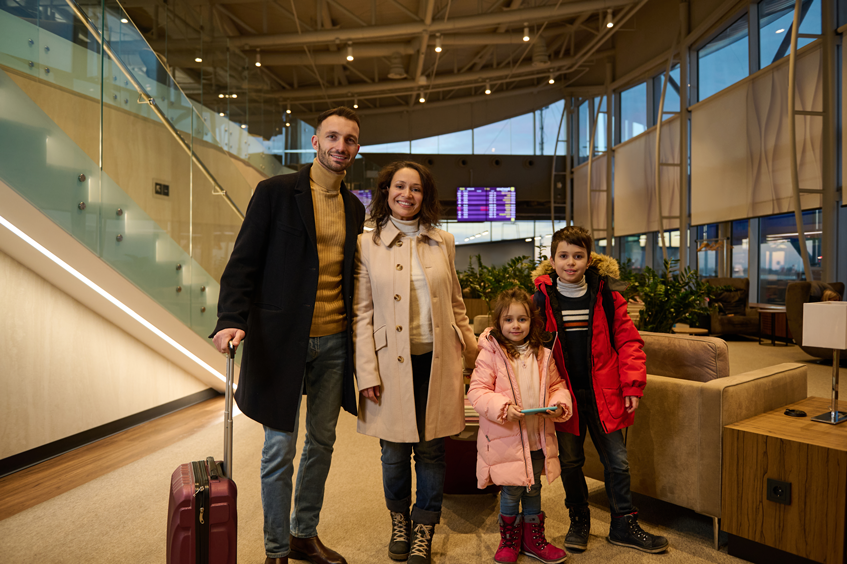 happy-family-enjoying-trip-together-stands-with-luggage-lobby-airport-departure-terminal-waiting-boarding-background-information-panel-with-flight-schedule-2 Family arriving at the airport, representing the success of spousal sponsorship in Canada.