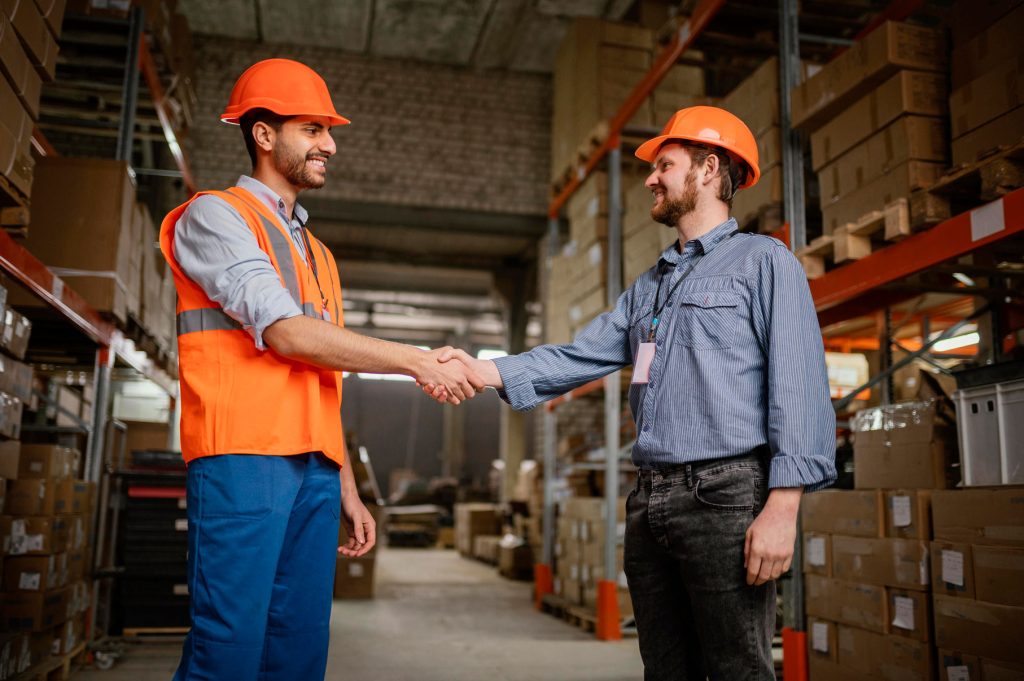 men-safety-equipment-work-2 Two workers in safety gear shaking hands in a warehouse, representing successful LMIA-based employment in Canada.