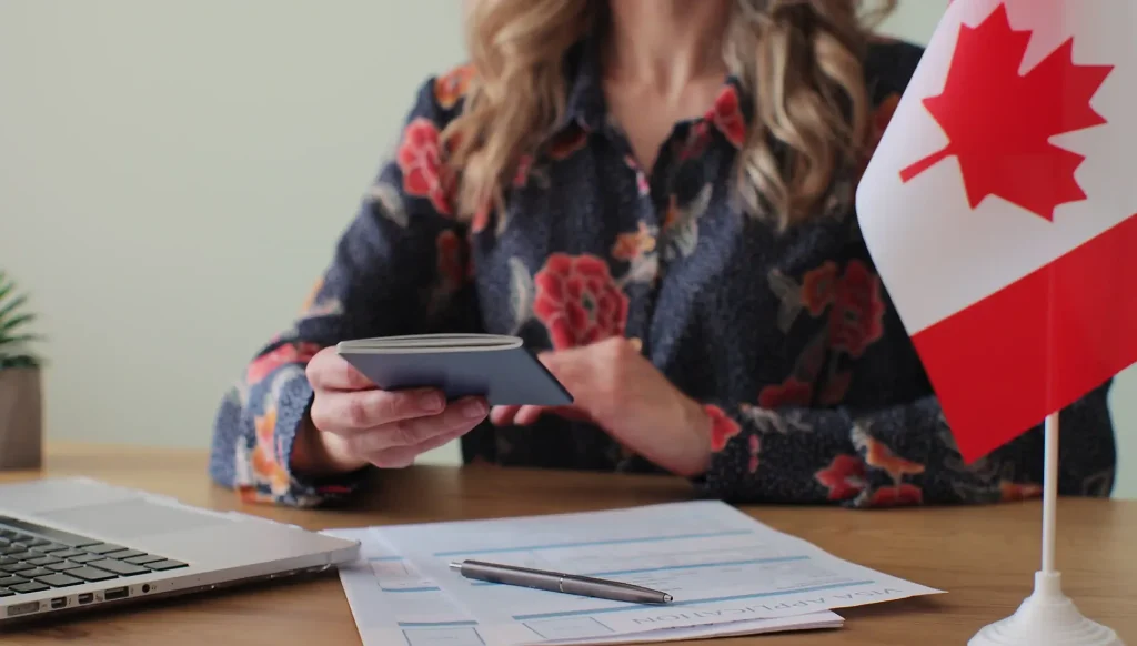 Woman holding a passport next to Canadian immigration forms and a flag, representing application readiness.