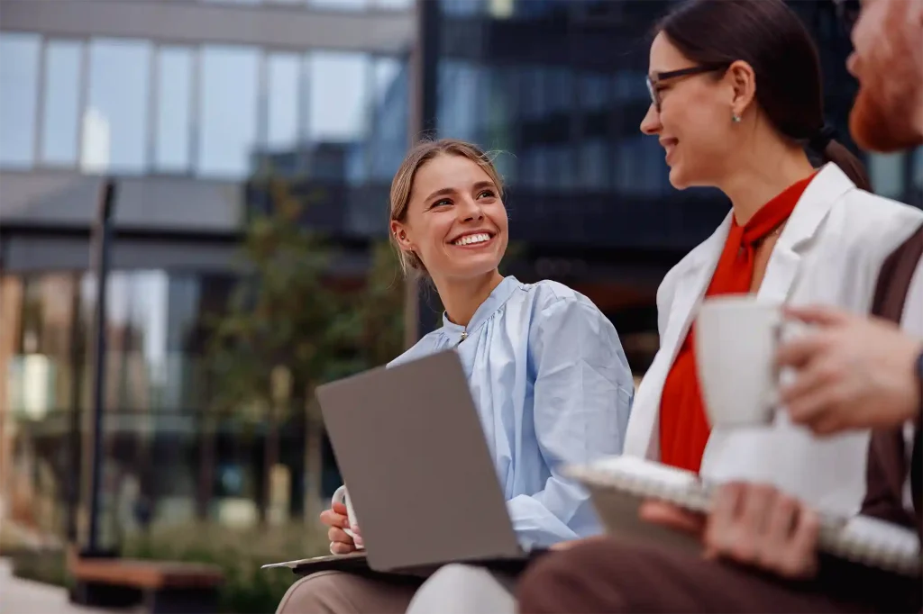 Smiling business professionals discussing immigration plans outdoors with laptops and coffee, reflecting friendly client support.